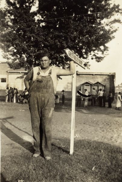 A young man in overalls standing next to a Green Valley Dairy sign.  Scene from the Lumberton textile workers strike.