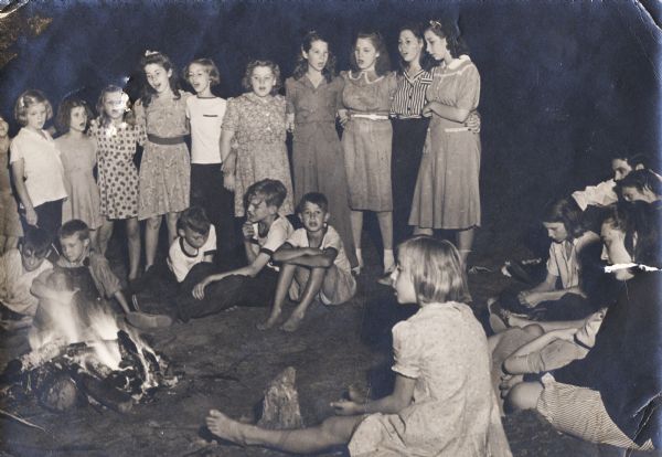 A group of girls and boys singing near a campfire, possibly part of Highlander Children's Camp.