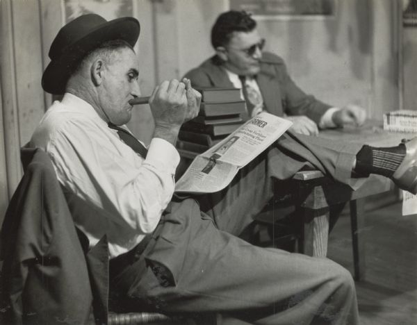 Two men smoking cigars, one in the process of lighting his while reading a newspaper, possibly the "Union Farmer".
