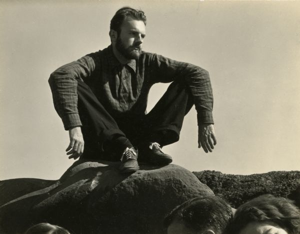 Portrait of a bearded man sitting on a rock. The tops of the heads of three people are visible in the lower foreground.