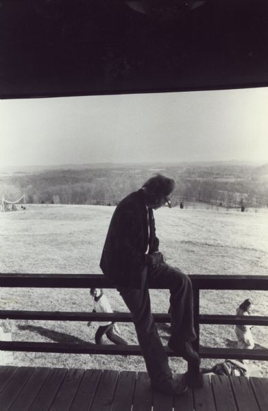 Rear view of an aging Myles Horton, co-founder of Highlander Folk School, peering over the side of a raised porch with a view to the horizon. There are two women on the lawn below the porch.
