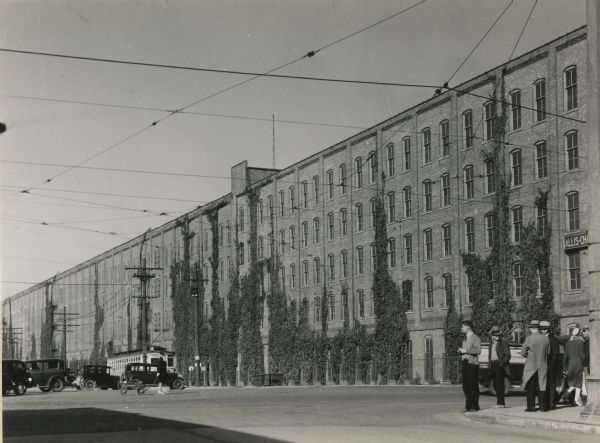 Exterior of factory with a streetcar and parked cars in the background. Streetcar wires crisscross overhead, and in the foreground on the right are a group of people standing at the corner of the road, while cars go past on the left.  Wall of building is also covered in several places with climbing ivy.