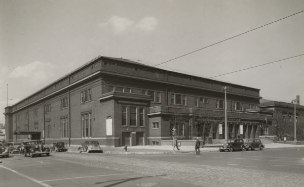 Exterior view of the building. Image has several cars and two people in front.