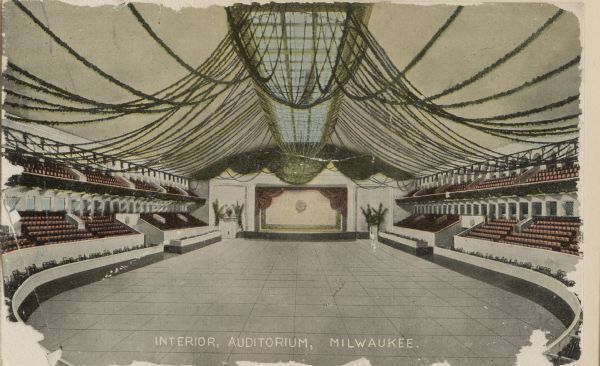 Interior of auditorium with a large open dance floor, a stage in the center background, and seats on the sides with balcony seating. The ceiling has a long window that has streamers reaching from it to the walls. Caption reads: "Interior, Auditorium, Milwaukee."