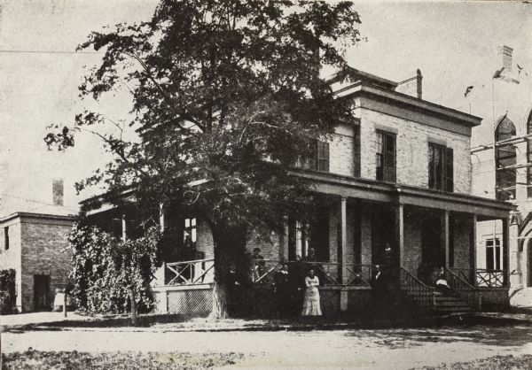 Large house with tree and several people on the porch and in front of the house. The building served as the Milwaukee Hospital in 1863.