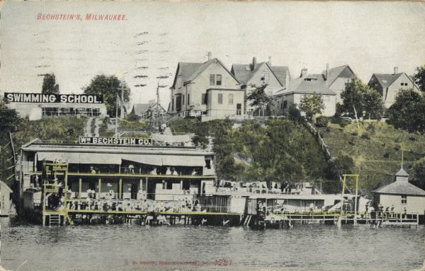 Color postcard of a bathing place on the water. A hill is behind the building, and there are a signs for a swimming school and the Wm. Bechstein Co. Several houses are along the top of the hill on the right. Caption reads: "Bechstein's, Milwaukee."