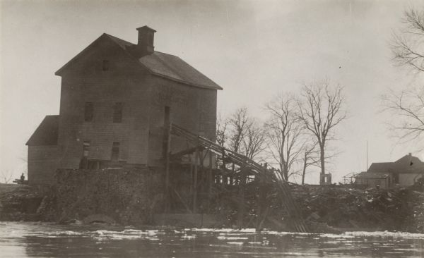 Mill on the Milwaukee River, with some bare trees and various objects on the riverbank.  The mill was destroyed in a flood in August of 1924.