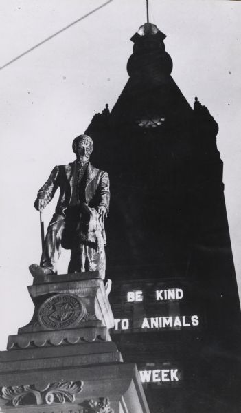 Statue with City Hall behind. Building has large neon letters that say "Be Kind to Animals Week". Monument is a memorial by John H. Mahoney to Henry Bergh, the founder of the American Humane Society. It was dedicated in 1891.