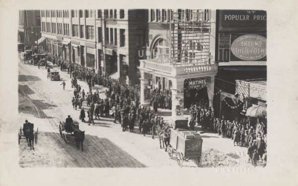 Theater with many signs, one of which is advertising a matinee.  Several other buildings are visible to the left of the building.  A large crowd of people line the street along the buildings, attempting to enter the theater. Several horse-drawn vehicles pass by on the dirt road.