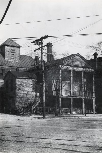 House from the east elevation.  Part of Historic American Buildings.  Image has a brick road, telephone poles and wire, and a building in the back with a sign reading "P. Schmitz & Son".  House has a roofed porch on the second level.