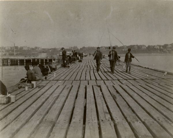 Long wooden pier with people fishing along the pier.  Three males with their fishing poles and buckets are walking toward the viewer.  A person next to a bike stands behind them.