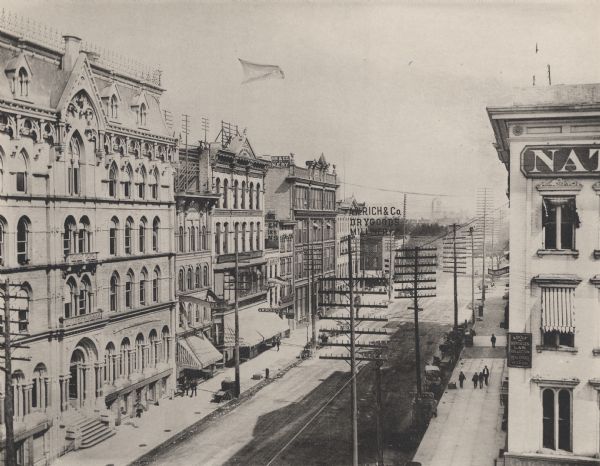 Elevated view of Broadway with several buildings lining the paved road, including A.W. Rich & Co. Drygoods, Millinery, and a Law Office on the right.  Large telephone or power line poles run along the street, and pedestrians can be seen on the sidewalks.