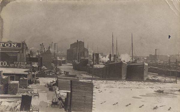 Two ships at the end of the canal, several buildings, a small lumberyard, and snow. Three other ships are visible down the canal.