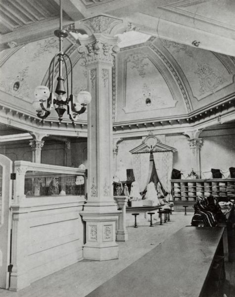 Interior view with dome visible. A long counter takes up the lower right of the image, and at the left is a white barrier wall, next to a decorated column.  Various goods can be seen on the counter in the back of the image.  A small chandelier hangs from the ceiling.