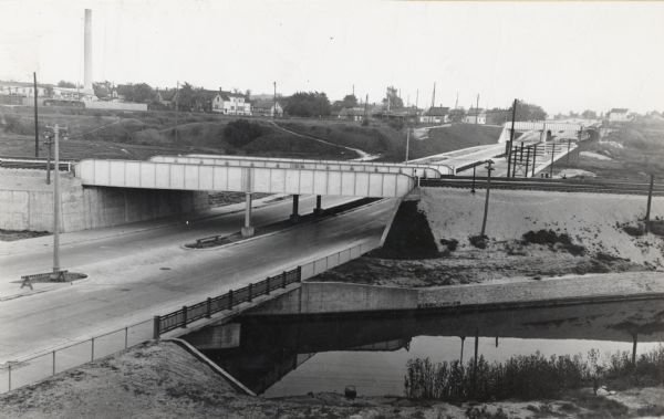 Looking south.  Bridge in foreground is over Kinnickinnic River.  Bridge over the road holds railroad tracks.  In background are several houses and a building with a large smokestack.  Telephone poles run along the road and among the houses.