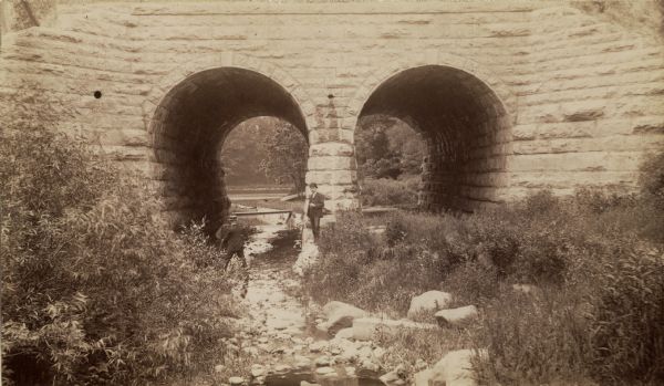 Chicago & North Western Railway viaduct on the line along the Milwaukee River.  The viaduct has two arched passages, and stones and rocks lining the path for the water.  Two men stand near the viaduct.  One man has a camera and is facing the stonework, the other man is looking out at the viewer.  Beyond the viaduct is more water and a grassy area with trees; low shrub and plant life are in the foreground.