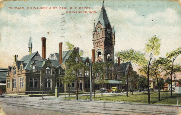 Angled view across street toward a park on the right and the railroad station behind. The station has blue roofs, chimneys, and a clocktower. Several vehicles are parked near the entrance. Caption reads: ", Milwaukee."Caption reads: "Chicago, Milwaukee, & St. Paul R.R. Depot, Milwaukee, Wis."