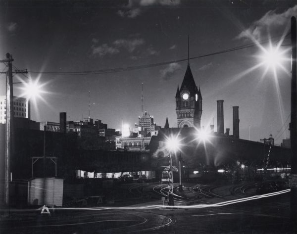 Nighttime image of the Depot and yard. Back of station, tracks, and a couple of trains are visible, as well as some of the buildings of Milwaukee in the background. Headlights are visible due to the long camera exposure.