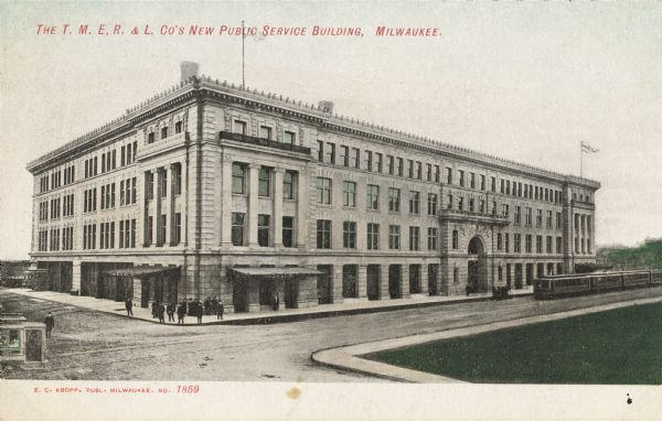 Slightly elevated view across street towards the building on a corner. Flags are on the roof on either end. A trolley is coming in from the right, and a groups of people is on the sidewalk at the corner. Caption reads: "The T.M.E.R. & L. Co's New Public Service Building, Milwaukee."