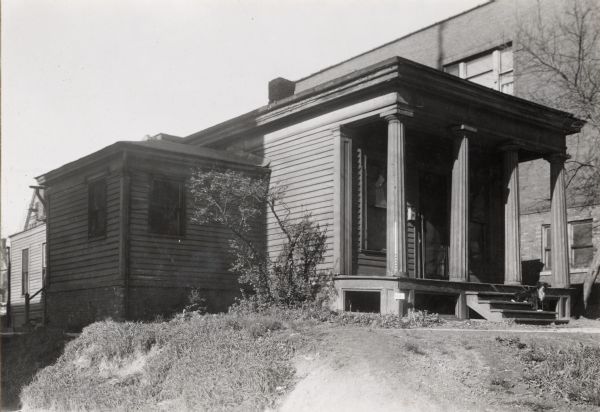 View from the southeast. House is one story, with a larger building behind it.  The porch has four fluted columns and siding along the walls.  A side room on the left of the image is in shadow, while the rest of the building is in the light.  A dirt path leads up to the image, and a plant grows in the corner of the yard next to the house.