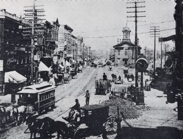 Elevated view of old City Hall building at right center facing the camera. Electric power poles are along the street, along which horse-drawn carriages and trolleys travel. A large clock is on the sidewalk to the right, and many buildings line the road into the distance.
