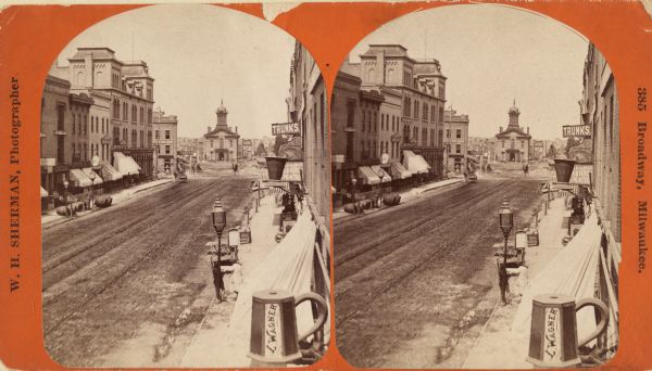 Stereograph; view of the earlier City Hall building, on Market Square at the corner of E. Water Street and Oneida Street. Several signs are in the image, including one in the lower right shaped like a mug with "L. Wagner" on the sde. Snow lies on the sidewalk and on the overhangs in front of stores.  Two people are standing next to a lamp post in the lower right, and a number of large barrels are on the sidewalk on the left.