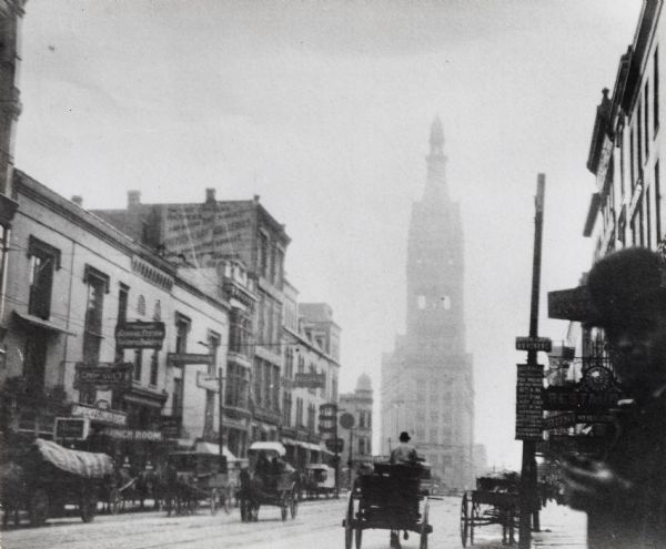 Street view; City Hall is in background.  Taken on the edge of a street with horse-drawn carriages, and buildings lining the street, and a man wearing a hat in the bottom right corner.