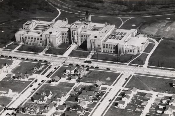 Aerial view of High School, in the upper half of the image, with fields, trees, and roadways surrounding it.  In the lower half is a residential neighborhood.