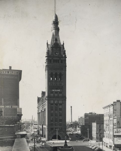 View towards the front of City Hall, taken from a rooftop. Streets, cars, a statue and a fountain are around the building. The sign on City Hall reads: "Welcome."