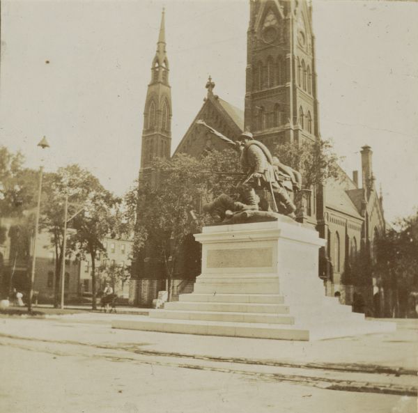 View of monument from the sidewalk, with a church behind. Several trees and lampposts are visible.