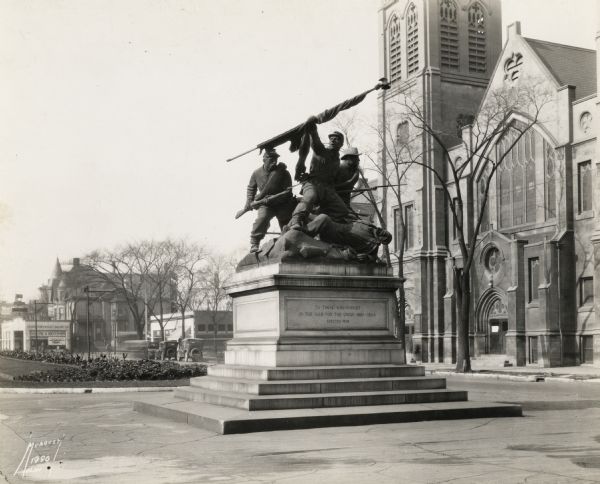 The Court of Honor, Wisconsin Avenue. The statue, "The Victorious Charge," is in the center, with a church behind. On the left are more buildings going into the distance, with cars and park area.  On the statue is inscribed "To Those Who Fought In The War For The Union 1861-1865. Erected 1958."