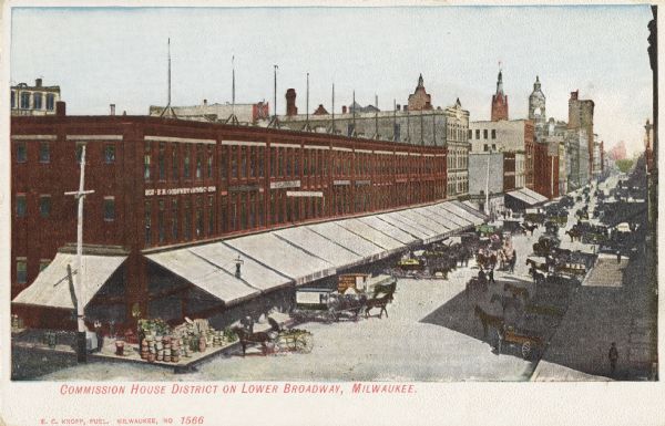 Elevated view of Lower Broadway with horse-drawn vehicles on both sides of the road. A long red building is in the left foreground angling to the right. A road runs alongside buildings with many horse-drawn vehicles. Caption reads: "Commission House District on Lower Broadway, Milwaukee."
