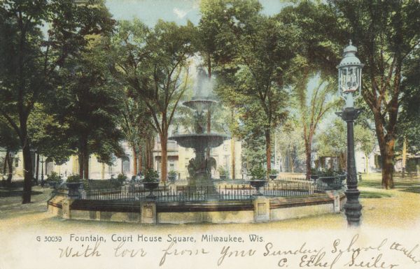Fountain surrounded by a path and trees. A lamppost is in the right foreground and buildings are in the background. Caption reads: "Fountain, Court House Square, Milwaukee, Wis."