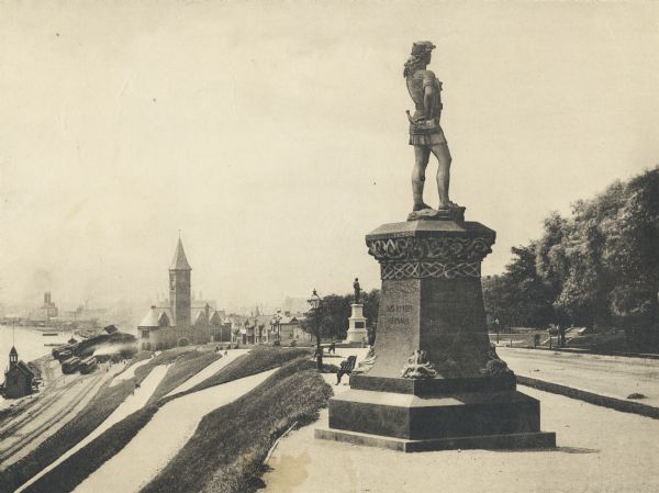 Side view of the statue on a plinth with Celtic designs, with his right hand holding a horn resting on his hip. The statue is located in Juneau Park. On the right of the statue are trees and a road. Cityscape is in the background.  Railroad tracks and trains at a station are next to water on the left side.