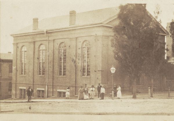 Church located at the corner of Broadway and Division Streets. A group of men, women and children stand at the corner near a large lamp post.