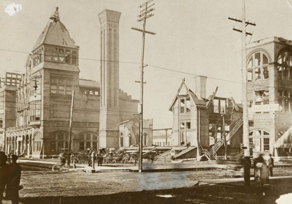 Afermath of the fire of June 4, 1905.  A chimney and some burnt out walls still stand.  The area around the ruins is fenced off.  The top of the building to the right shows some damage.  Some people are on the street corner next to the ruins, and across the street.  A sign for Ice Cream Soda is on the right.