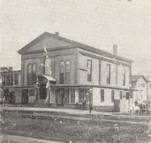At the corner of Wisconsin and Milwaukee Streets; the present site of T.A. Chapman Store.  View of building from across the street.  Three men stand in front of a window near the entrance.  A horse-drawn carriage is on the road at the right.  A lamp post stands at the corner.