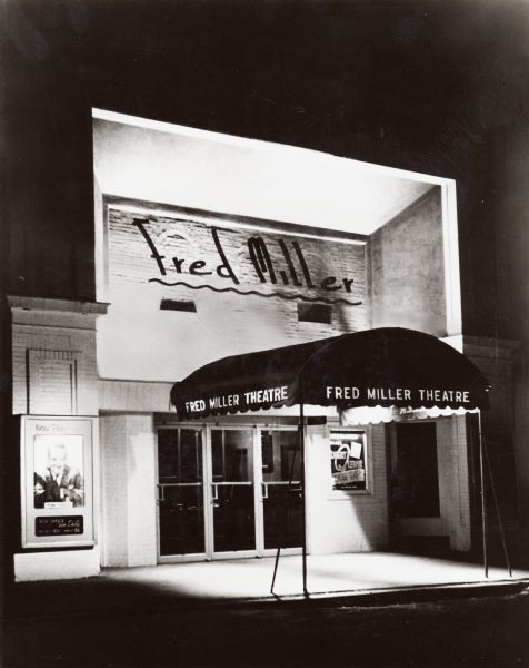 Nighttime view of theater entrance.  Doorway has a fabric overhang with the theatre name printed on each side.  Posters for Robert Lewis and Tom Daily flank the doors.