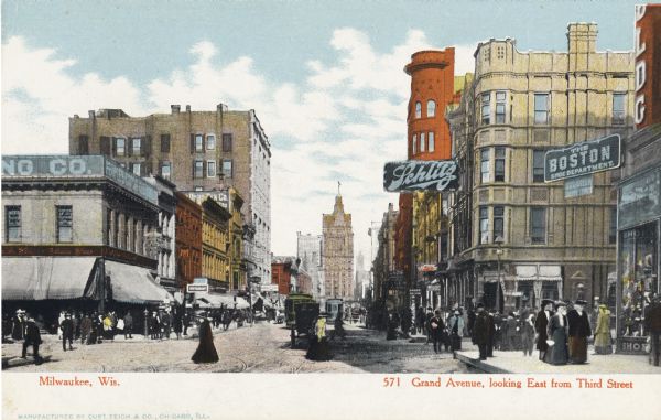 View looking east from Third Street. Buildings line the road, with signs and advertisements. Roads and sidewalks are crowded with horse-drawn vehicles and pedestrians. Captions reads: Caption read: "Milwaukee, Wis." and "Grand Avenue looking East from Third Street."