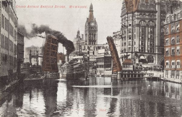 View along the river, with a raised bridge with a steamboat passing through. The river lined by buildings on both sides. A clocktower is in the background. Caption reads: "Grand Avenue Bascule Bridge, Milwaukee."
