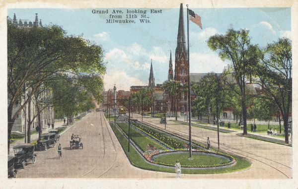Elevated view looking east from Eleventh Street. The avenue is lined with buildings and trees. A large flagpole is in the center of the avenue which is landscaped with flowers. Caption reads: "Grand Ave. looking East from 11th St., Milwaukee, Wis."