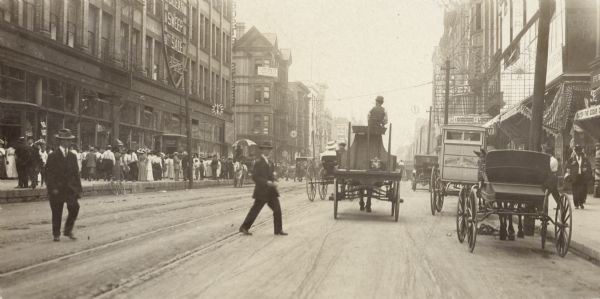 View down the avenue. A large crowd of people on are on the left in front of a storefront, and several horse-drawn vehicles are on the right.  Two men are crossing the road.