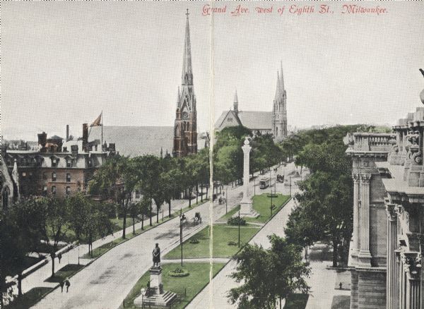 Elevated view of avenue west of Eight Street. On the left are two churches, a few other buildings, and trees. The road is split with a strip of land that has two monuments. Several horse-drawn vehicles are on the road. Caption reads: "Grand Ave. west of Eighth St., Milwaukee."