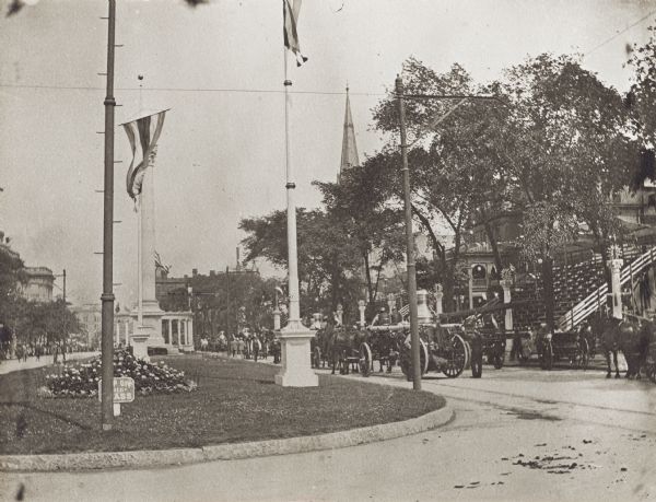 Flagpole on a strip of grass in the middle of an avenue. On the right are several horse-drawn vehicles and people parading in the road in front of what appears to be a grandstand.