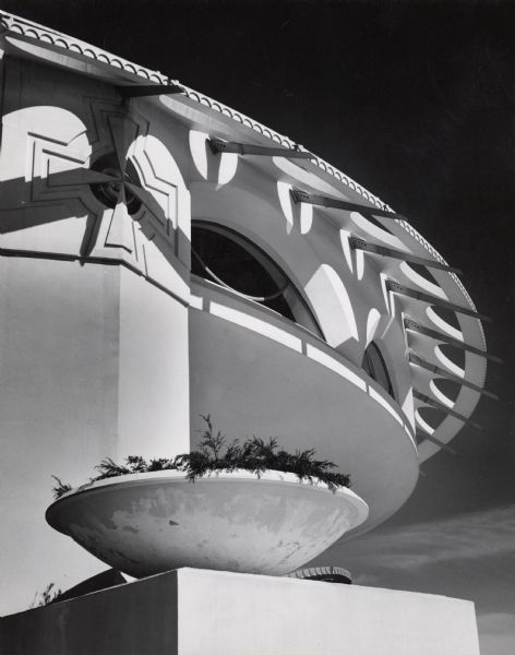 Partial view of the building, designed by Frank Lloyd Wright.  On the left is a cross on a pillar section, with a plant bowl on top of a block in front.  Arched windows top the walls under the roof.  The circular roof comes out several feet above the walls, decorated with metal supports and semi-circles.