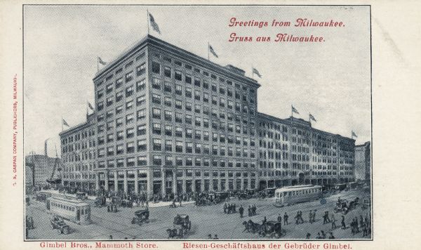 Elevated view toward the building on a street corner. The intersection in front is crowded with pedestrians, horse-drawn vehicles, and trolleys. Nine flags are at various points on the roof of the building. Caption at top reads: "Greetings from Milwaukee," "Gruss aus Milwaukee," and at bottom: "Gimbel Bros., Mammoth Store." and "Riesen-Geschaftshaus der Gebruder Gimbel.".