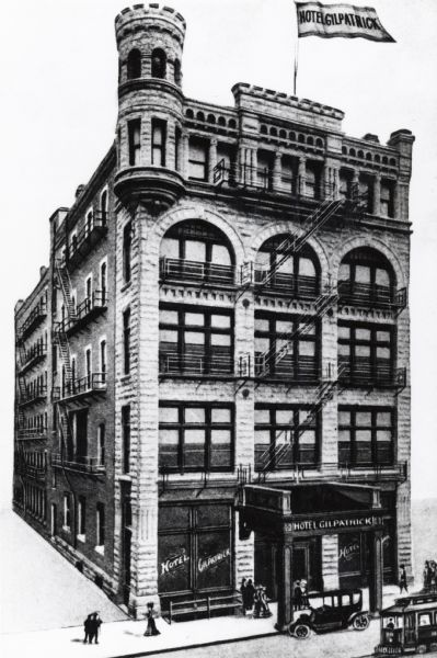 Five story building with a flag with the hotel's name on it flying from the roof.  Several people, a car, and a trolley are in front.