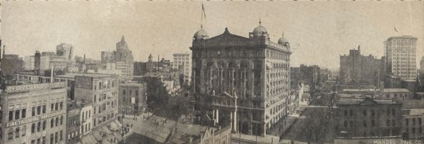 Panoramic view from the top of a building. Home of Merchants and Manufacturers' Association of Milwaukee. Building is in the center of the image, with two roads on either side. Buildings on the left have signs for Peoples Tailoring Co. and Mandel Engraving Company.