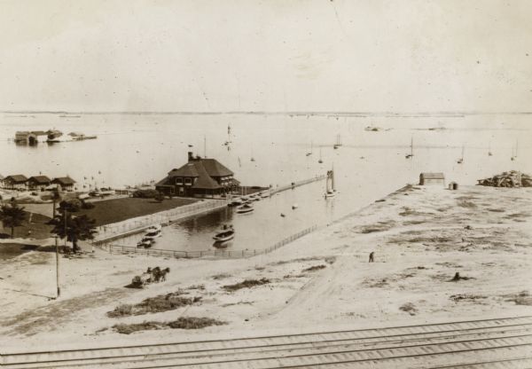 View from across railroad tracks.  Resort is on the water's edge, with a small inlet on the right.  Land is mostly sand, though a small part on the left has grass. Several boats are in the water.  A man drives a horse-drawn vehicle not far from the inlet.