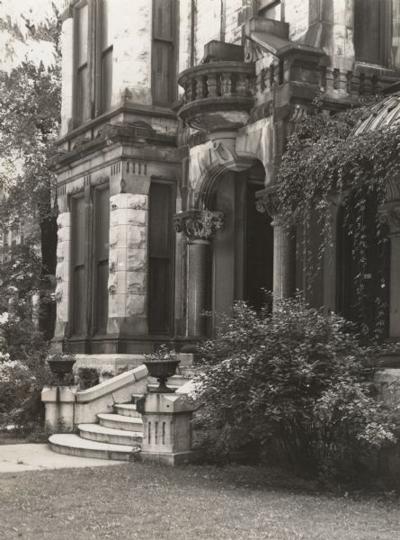 Entrance to large home. Part of porch as well as side of house visible.  Curved stairs lead up to the porch. Door flanked by columns with highly detailed capitals and lintel. Ivy grows along the slanted roof of the porch, and shrubbery grows in front in the lawn. House  is made of stone.